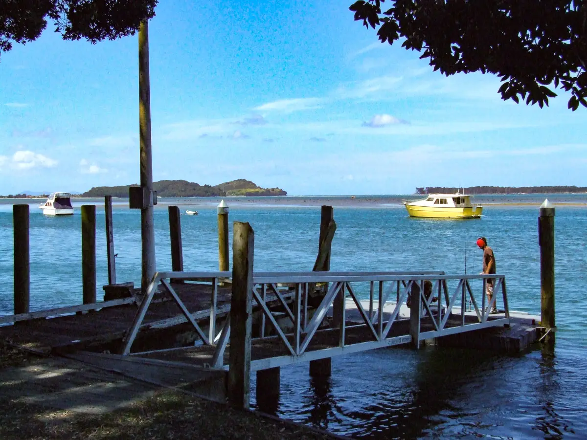 Person fishing off the Tanners Point wharf with boats moored in the Tuapiro Channel and Bowentown headland in the distance
