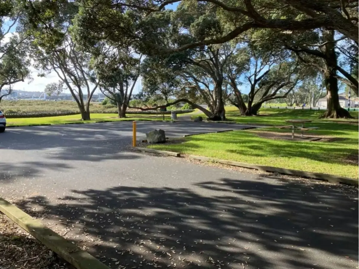 Shaded carpark under mature pohutukawa trees at Kiwi Parade Mangere Bridge with harbour views in background